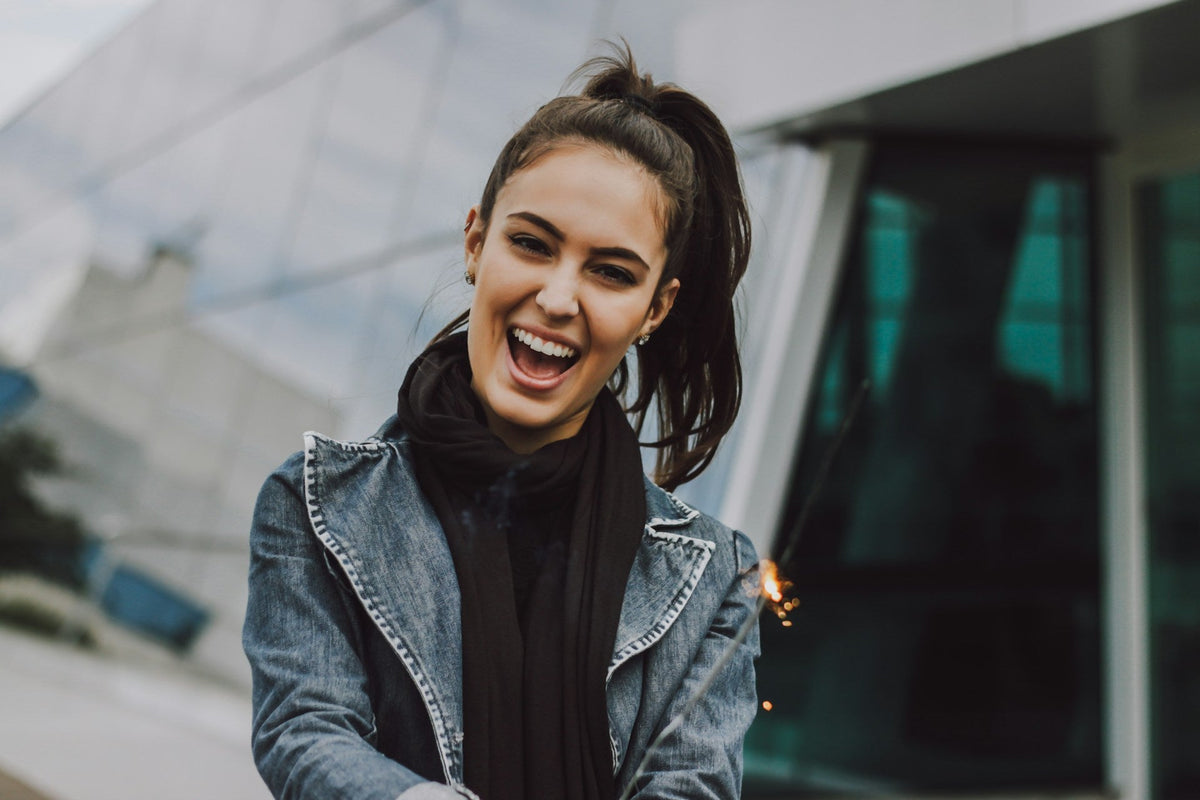 A laughing woman with a high ponytail, wearing a denim jacket and scarf, holding a sparkler.
